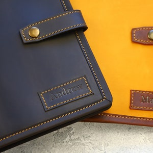 May include: Close-up of two personalized leather journals. One is dark brown with "Andrew" embossed, the other is yellow with "Maria" embossed. Both have brown straps with brass button closures and contrasting stitching.