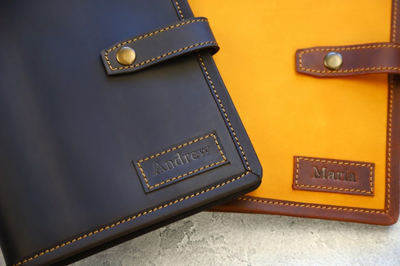 May include: Close-up of two personalized leather journals. One is dark brown with "Andrew" embossed, the other is yellow with "Maria" embossed. Both have brown straps with brass button closures and contrasting stitching.