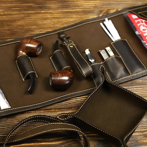 Peut inclure: Un étui en cuir marron roulé avec plusieurs poches et compartiments. L'étui contient deux pipes, un petit outil et une petite pochette. L'étui est ouvert et posé à plat sur une surface en bois.