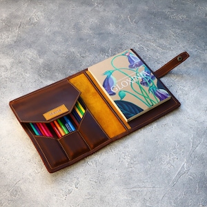 May include: Open leather pencil case with a notebook. The case is brown with a yellow interior and a flap closure. The notebook has a floral design and the word "GLOXINIA" on the cover. A variety of colored pencils are visible.