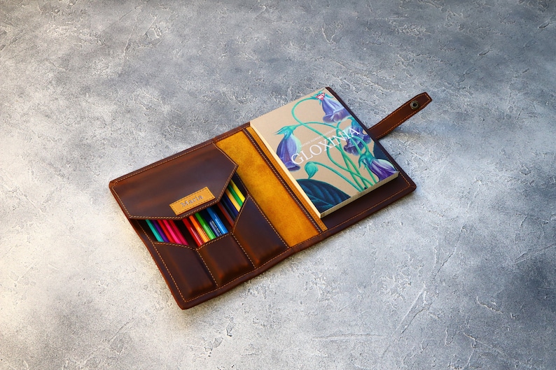 May include: Open leather pencil case with a notebook. The case is brown with a yellow interior and a flap closure. The notebook has a floral design and the word "GLOXINIA" on the cover. A variety of colored pencils are visible.