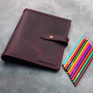 May include: A burgundy leather portfolio with a flap closure and a brass button. The portfolio has the word "Melissa" embossed on the front. A set of colored pencils are lying on a gray surface next to the portfolio.