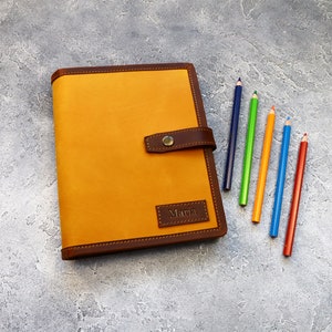 May include: A mustard-colored leather journal with a dark brown border and clasp. The journal has the name "Maria" embossed on a small rectangular patch. Six colored pencils in blue, green, orange, red, and yellow are arranged next to the journal.