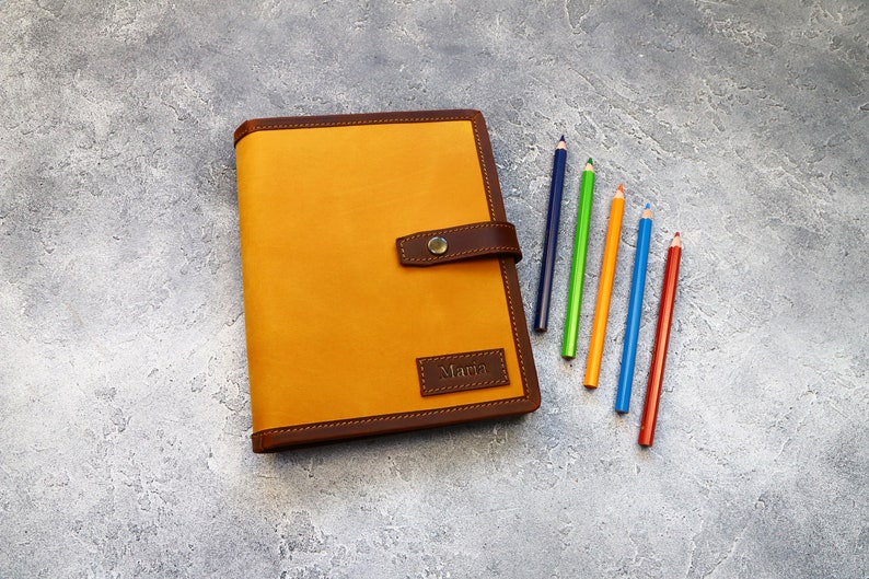 May include: A mustard-colored leather journal with a dark brown border and clasp. The journal has the name "Maria" embossed on a small rectangular patch. Six colored pencils in blue, green, orange, red, and yellow are arranged next to the journal.