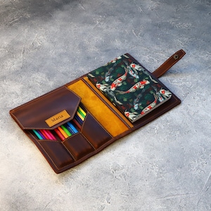 May include: A brown leather pencil case with a flap closure, containing colored pencils. A small notebook with a koi fish pattern is visible. The case has a yellow interior and a small label that says "Maria".