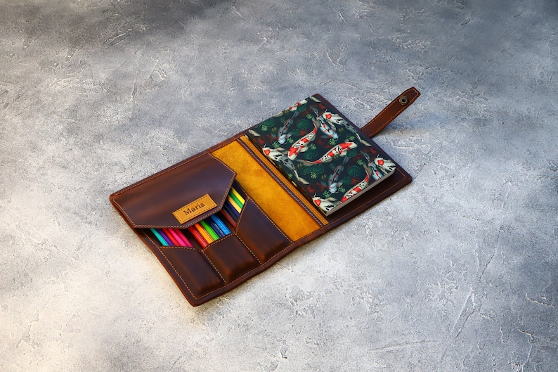 May include: A brown leather pencil case with a flap closure, containing colored pencils. A small notebook with a koi fish pattern is visible. The case has a yellow interior and a small label that says "Maria".