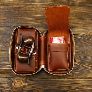 May include: A brown leather carrying case with a zipper closure. The case is open and contains two wooden pipes and a small red pouch. The case is on a wooden surface.