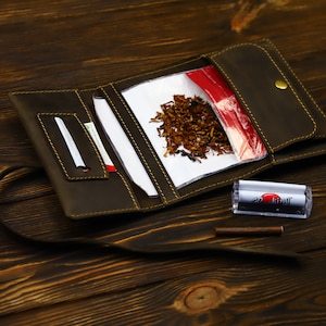 Puede incluir: Una bandeja de enrollado de cuero marrón con una solapa que se abre para revelar una bolsa de plástico transparente con tabaco y una bolsa de plástico roja. También se puede ver una máquina de enrollar de plástico transparente y un palo de enrollar de madera marrón.