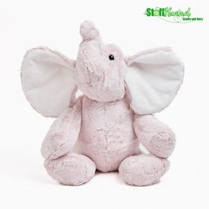 Könnte beinhalten: Ein Plüschelefant mit weichem, hellrosa Fell und großen, glatten weißen Innenohren. Das Stofftier hat einen nach oben gerichteten Rüssel, kleine schwarze Augen und eine sitzende Haltung. Das Logo StoffHummel Stoffe mit Herz ist zu sehen.