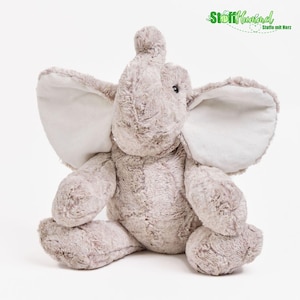 May include: A plush elephant toy made of soft, light brown, textured faux fur. The stuffed animal features large, floppy ears with smooth, cream-colored inner linings, a raised trunk, and dark, round eyes. The logo StoffHummel Stoffe mit Herz is visible.