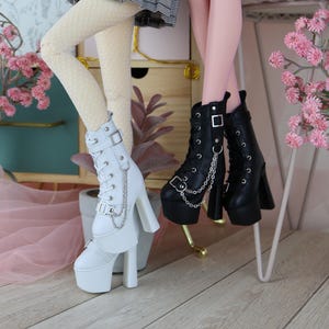 May include: Two dolls wearing platform boots. One doll wears white boots with silver chain and buckle details. The other doll wears black boots with silver chain and buckle details. The dolls are wearing skirts and tights.