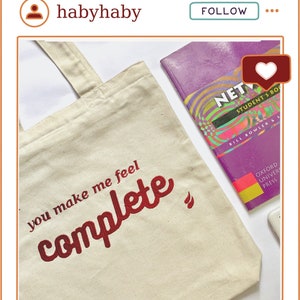 May include: A white canvas tote bag with the text "you make me feel complete" in red embroidery. The bag is lying on a table next to a purple book with the title "NET" and a white phone.