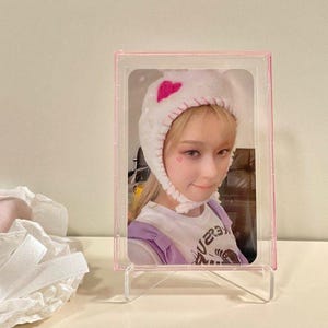 May include: A rectangular pink plastic card holder frame containing a photograph of a person wearing a white fuzzy bonnet with a pink heart detail. The person has a small pink heart mark on their cheek and wears a white t-shirt with purple straps.