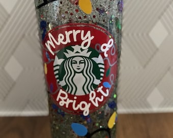 Może przedstawiać: Przezroczysty kubek z zielonym, czerwonym i złotym brokatem. Kubek ma czarno-białe logo Starbucks z napisem "Merry & Bright" w kolorze czerwonym i białym.
