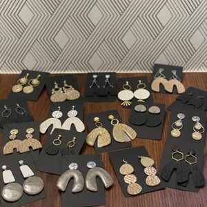 May include: Assortment of handmade polymer clay earrings in various shapes, colors, and patterns. The earrings are displayed on black card stock with white backing. Some earrings feature geometric shapes, while others have a more organic, free-flowing design.