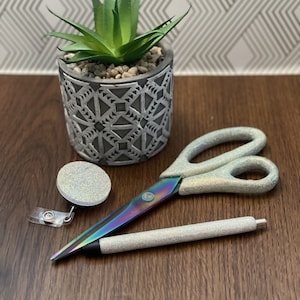 Puede incluir: Un conjunto de artículos de oficina con purpurina iridiscente que incluye un portagajes retráctil, un bolígrafo y unas tijeras. Los artículos están sobre una superficie de madera junto a una pequeña planta suculenta en maceta.