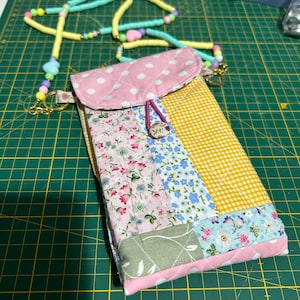 May include: A patchwork fabric phone case with a pink polka dot flap and a yellow gingham panel. The case is adorned with floral and patterned fabric pieces. A beaded lanyard in pastel colors is attached.