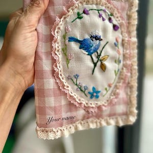 以下が含まれることがあります： ピンクと白のギンガムチェックの生地のポーチで、レースのトリムと刺繍された青い鳥と花が施されています。ポーチには、下部に「Your name」という刺繍が施されています。