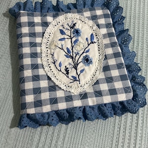 以下が含まれることがあります： 青と白のギンガムチェックの生地のポーチ。白いレースの縁取りと、白い楕円形に青い花が刺繍されている。