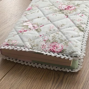 May include: A book cover with a floral pattern in shades of pink, green, and white. The cover is quilted and has a white crocheted trim.
