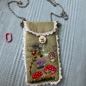 May include: Olive green crossbody phone bag with embroidered mushrooms, flowers, and foliage. The bag has a white lace trim and a silver chain strap. A small pink carabiner is attached. The bag is approximately 7 inches tall and 4 inches wide.