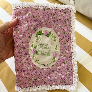May include: A pink fabric book cover with a white lace trim. The cover has a floral pattern and an embroidered oval with the words "Holy Bible" in cursive.