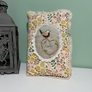 以下が含まれることがあります： 花柄とレースのトリムが付いた、クリーム色の小さな枕。枕には、鳥と「Mame」という文字が入ったハート型の刺繍のデザインが施されています。