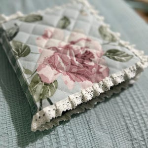May include: A quilted fabric pot holder with a floral pattern in shades of pink, green, and white. The pot holder has a white lace trim around the edges.