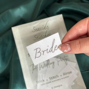 May include: A white label with the word "Bride" in brown script is held over a clear packet. The packet contains small white labels and text that reads "Seaside Stitches" and "The Wedding Party". The background is a dark green satin fabric.
