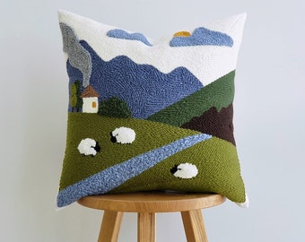 May include: A white pillow with a colorful embroidered landscape scene. The scene features a blue sky with clouds, green hills, a small house, and white sheep. The pillow is sitting on a wooden stool.