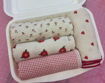 May include: A white clamshell container holding four rolled fabric cloths. One cloth features a small red floral pattern, another has larger red flowers, one has a red grid pattern, and the final cloth is cream with small red hearts.