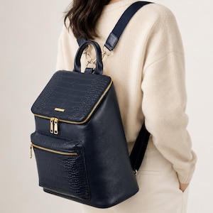 May include: A navy blue backpack with a faux crocodile skin texture on the top flap and front pocket. The backpack has gold-colored zippers and hardware, and a top handle. The straps are navy blue with a dark blue fabric.