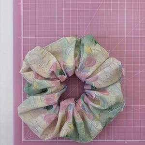 May include: A fabric hair scrunchie featuring a soft, painterly pattern of pastel pink tulips, muted green leaves, and yellow accents on a cream-coloured background. The scrunchie is positioned on a pink cutting mat with white grid lines.