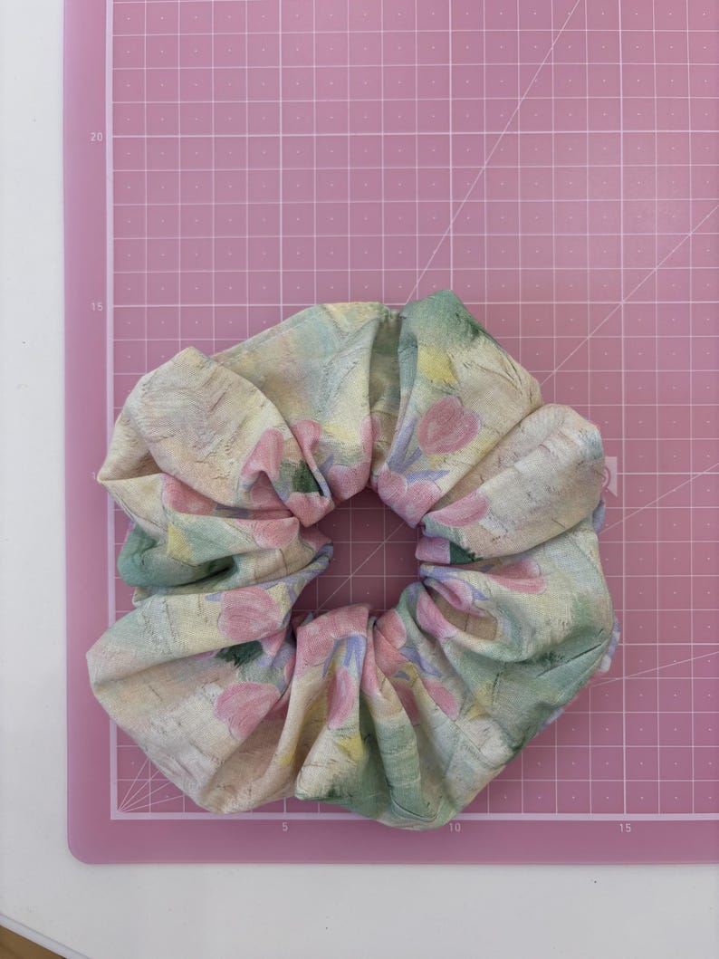 May include: A fabric hair scrunchie featuring a soft, painterly pattern of pastel pink tulips, muted green leaves, and yellow accents on a cream-coloured background. The scrunchie is positioned on a pink cutting mat with white grid lines.