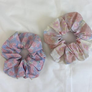 May include: Two fabric hair scrunchies. The left scrunchie features a light blue background with a repeating pattern of pink circular shapes. The right scrunchie has a white background with a repeating pattern of pink and yellow floral motifs.