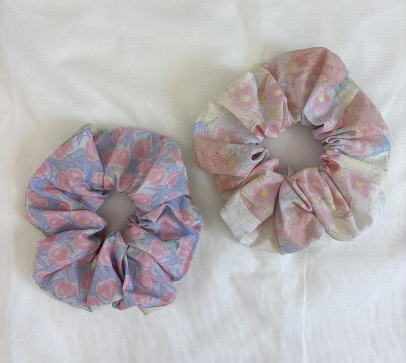 May include: Two fabric hair scrunchies. The left scrunchie features a light blue background with a repeating pattern of pink circular shapes. The right scrunchie has a white background with a repeating pattern of pink and yellow floral motifs.