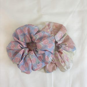 May include: Two fabric hair scrunchies featuring floral patterns. The left scrunchie has a light blue background with pink tulip-like flowers. The right scrunchie displays a soft, watercolour-style print with muted pink and cream floral motifs.