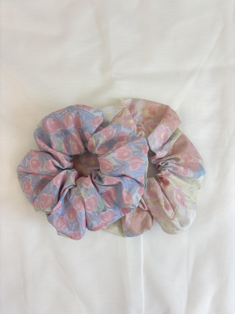 May include: Two fabric hair scrunchies featuring floral patterns. The left scrunchie has a light blue background with pink tulip-like flowers. The right scrunchie displays a soft, watercolour-style print with muted pink and cream floral motifs.