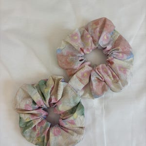 May include: Two fabric hair scrunchies featuring a soft watercolour-style floral pattern. The scrunchies display a palette of muted pink, pale green, and soft yellow tones on a light background, with gathered fabric creating a ruffled texture.