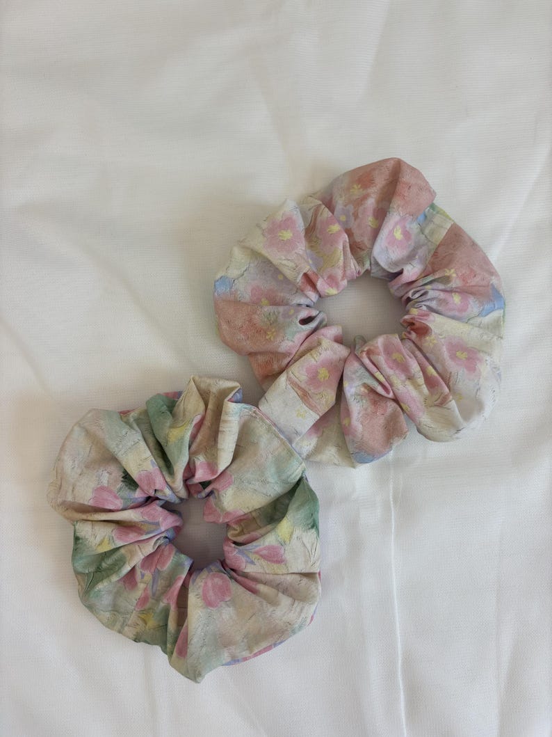 May include: Two fabric hair scrunchies featuring a soft watercolour-style floral pattern. The scrunchies display a palette of muted pink, pale green, and soft yellow tones on a light background, with gathered fabric creating a ruffled texture.