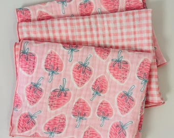 May include: A set of folded muslin blankets featuring a repeating pattern of pink strawberries with teal stems on a light pink background, layered with a second blanket displaying a classic pink and white gingham check pattern with finished edges.