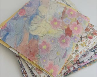May include: A stack of square fabric swatches featuring various floral and botanical patterns. The top swatch displays a soft, painterly design with pink, yellow, and blue flowers. Beneath it are layers of fabric with intricate, multi-coloured floral prints.