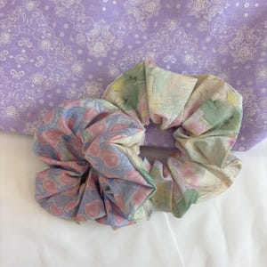 May include: Two fabric hair scrunchies. One scrunchie features a light blue background with a repeating pattern of pink peaches and green leaves. The second scrunchie has a watercolour-style abstract print in shades of green, yellow, and pink.