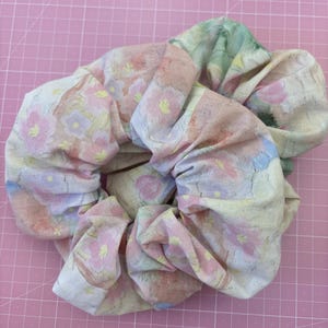 May include: A fabric hair scrunchie featuring a soft pastel floral pattern with shades of pink, yellow, blue, and green. The material has a textured, woven appearance with gathered folds that create a voluminous, circular shape for hair styling.
