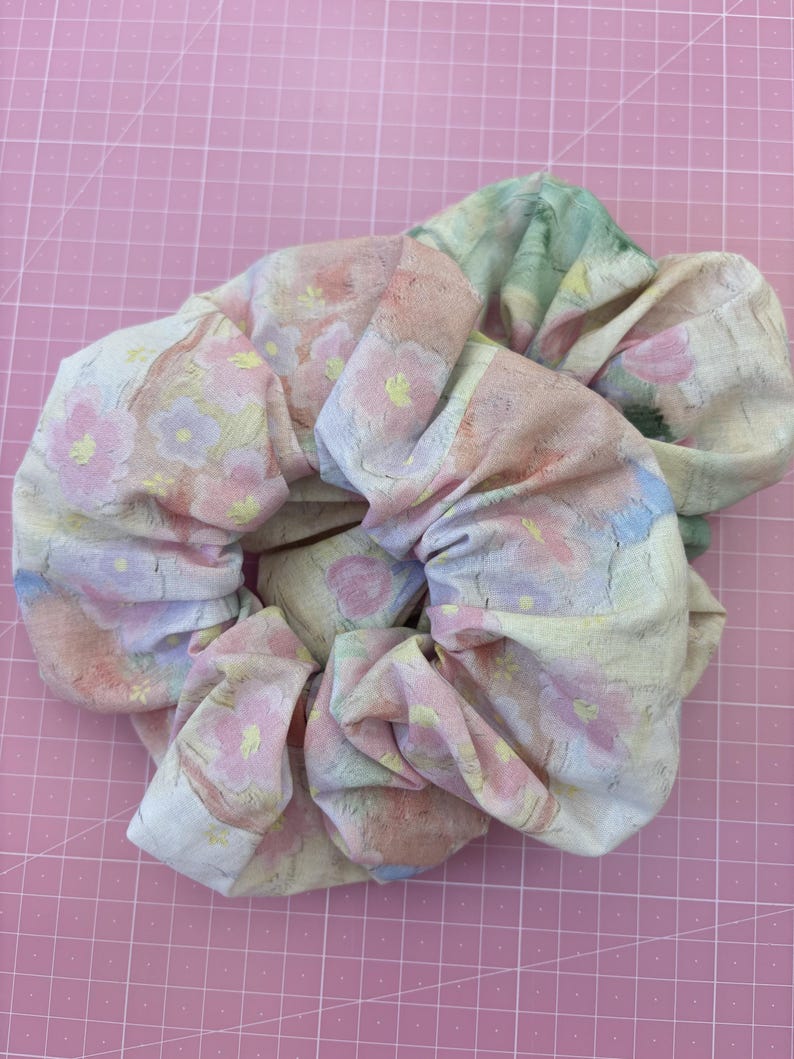 May include: A fabric hair scrunchie featuring a soft pastel floral pattern with shades of pink, yellow, blue, and green. The material has a textured, woven appearance with gathered folds that create a voluminous, circular shape for hair styling.