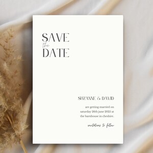 May include: A cream-colored Save the Date card with elegant typography. The text reads "SAVE the DATE" at the top, followed by the names "SUZANNE & DAVID" and wedding details. Dried ornamental grass is in the background.