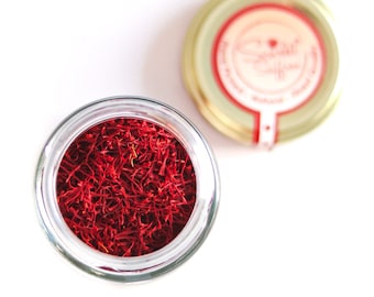 May include: A glass jar filled with dried red saffron threads. The jar has a clear glass lid with a gold colored metal rim.