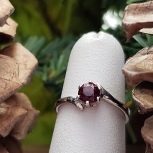May include: A silver ring with a dark red gemstone set in a prong setting. The ring has a curved band.