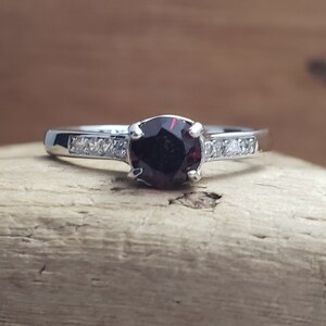 May include: A silver ring with a dark red gemstone surrounded by smaller clear gemstones.