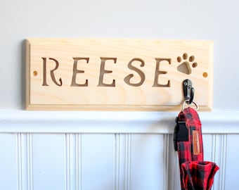 May include: A wooden sign with the name "REESE" carved into it. A paw print is carved next to the name. A black hook is attached to the sign, and a red and black checked dog collar is hanging from the hook.